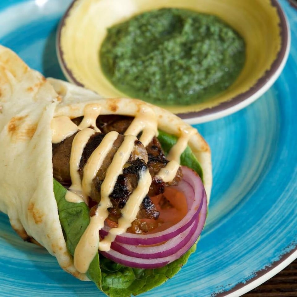 Grilled meat pita wrap with lettuce, red onion, creamy sauce, and green chutney on a blue plate.