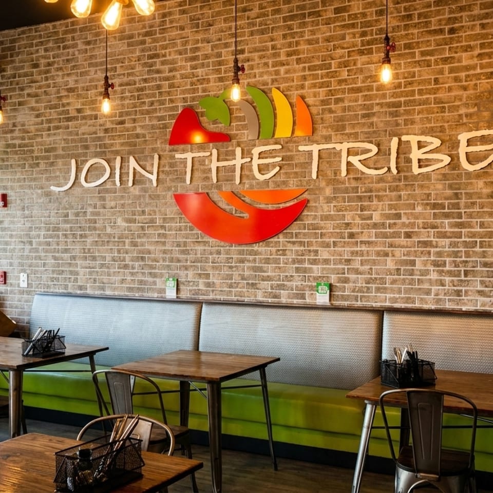 Restaurant interior with brick wall, "Join the Tribe" sign, green booth seating, and wooden tables.