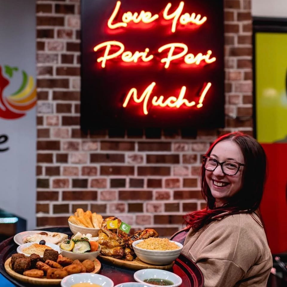 Smiling woman sits beside a table laden with assorted dishes and dipping sauces beneath a neon “We Love You Peri Peri Much!” sign.