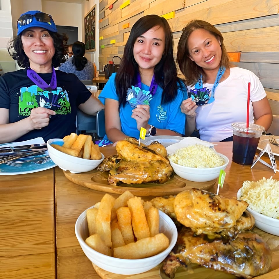 Three smiling women with marathon medals enjoy roasted chicken, fries, and rice at a restaurant.