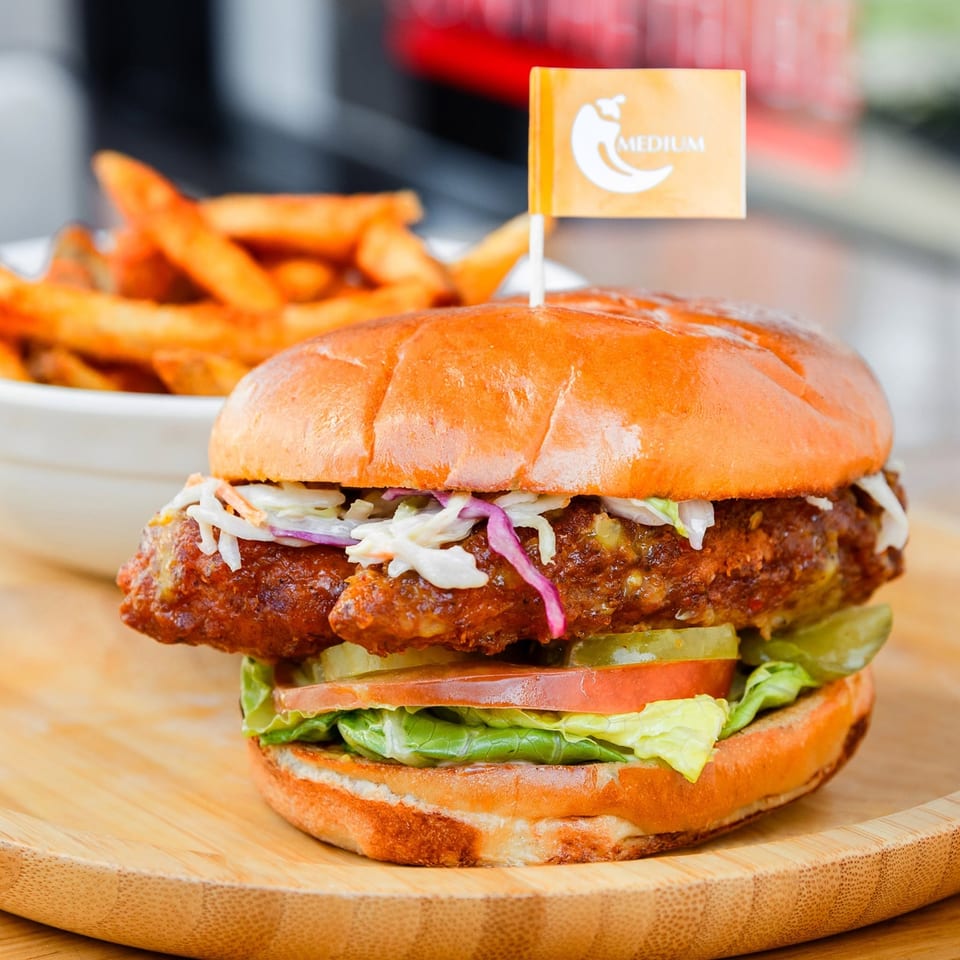 Medium spicy chicken sandwich with coleslaw, pickles, lettuce, tomato, and fries on a wooden plate.