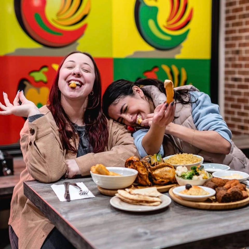 Two women laugh while sharing a colorful spread of grilled meats, rice, flatbreads, and side dishes at a restaurant table.